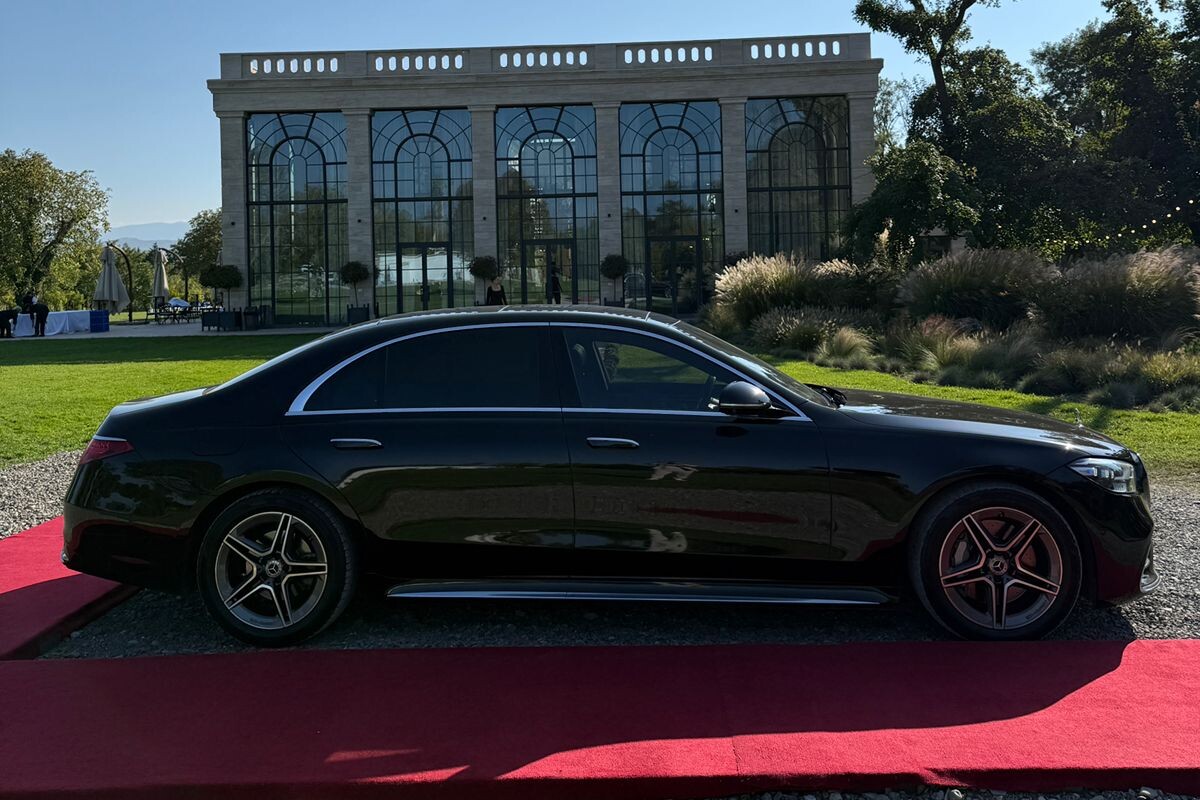 შავი Mercedes S-Class წითელ ხალიჩაზე გაჩერებული თბილისთან ახლოს, მინის პავილიონის თაღოვანი ფანჯრების ფონზე.