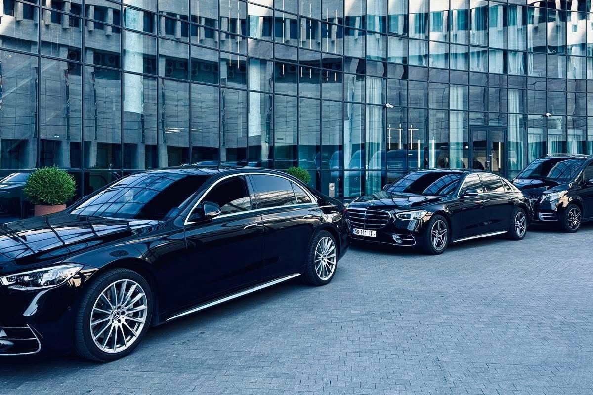 სამი შავი Mercedes-Benz სედანი რიგში დგას მოხრილი მინის საოფისე შენობის წინ, ნათელ დღეს.