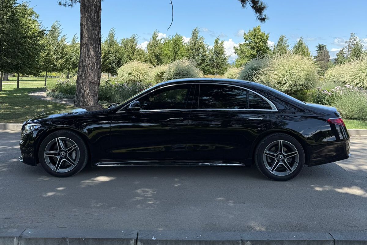 SOIT-ის ავტოპარკის შავი Mercedes S-Class გვერდიდან, პარკის გზაზე, ზაფხულში, ფონზე ხეები და აყვავებული ბუჩქები.