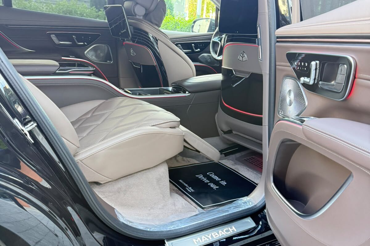 Mercedes-Maybach EQS 680-ის გაღებული უკანა კარი: ღია ფერის ნაკერიანი სავარძელი, ხალიჩა და MAYBACH ზღურბლი.