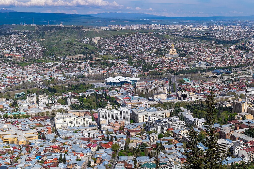ცენტრალური თბილისის ფართო ხედი, ქალაქი გადაშლილია მტკვრის ხეობაში, ახალი მაღალსართულიანი სასტუმროებითა და საცხოვრებელი კოშკებით, რომლებიც ძველი წითელ-სახურავიანი უბნების გვერდით ამოდის.