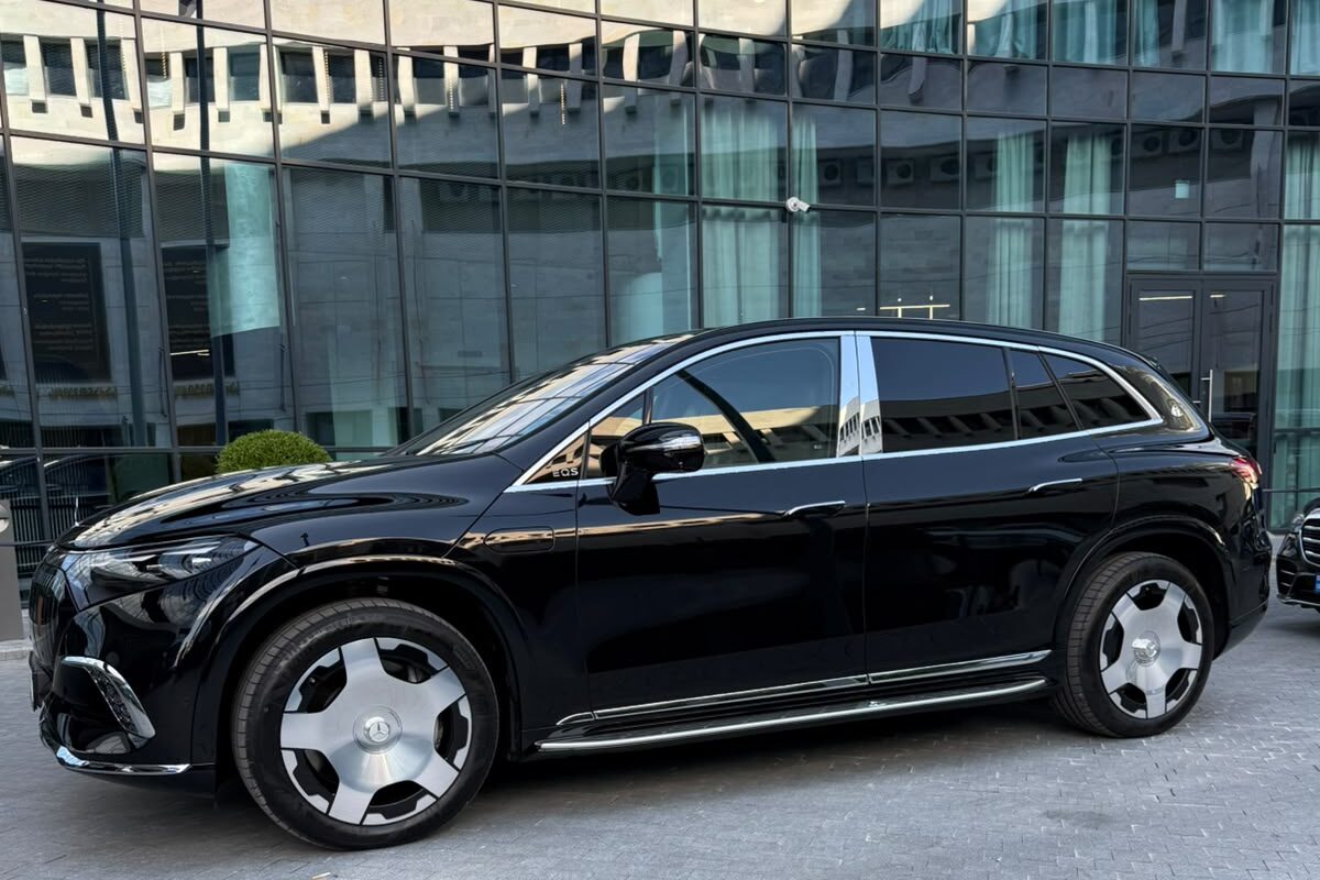 შავი Mercedes-Maybach EQS 680 გვერდიდან, ქვაფენილზე, შუშის ფასადის მქონე შენობის წინ, თბილისში.