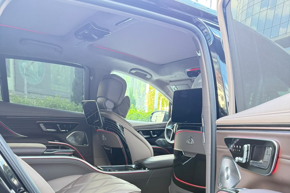 Mercedes-Maybach EQS 680-ის უკანა სალონი გაღებული კარით, ორი ცალკეული სავარძლით ღია ნაკერიან ტყავში და განათებული MAYBACH ზღურბლით.