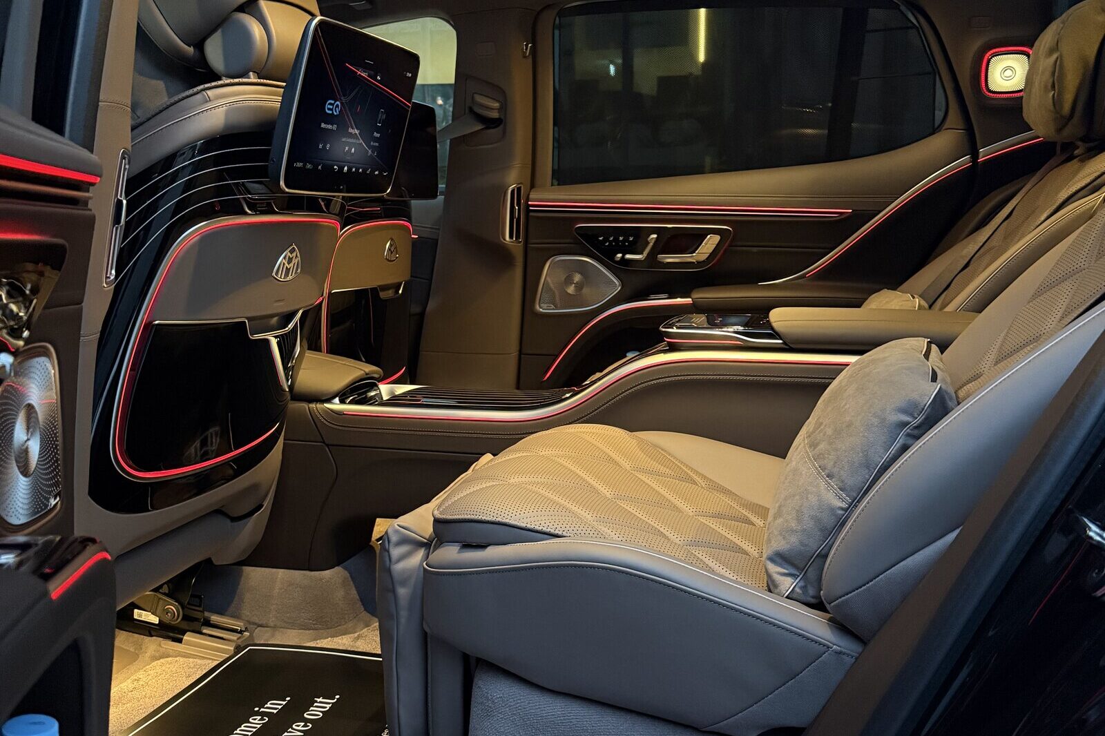 Mercedes-Maybach EQS-ის უკანა სალონი გაღებული მარცხენა კარიდან: ნაკერიანი სავარძელი დახრილი საზურგით, ბალიში და წინ ეკრანი.