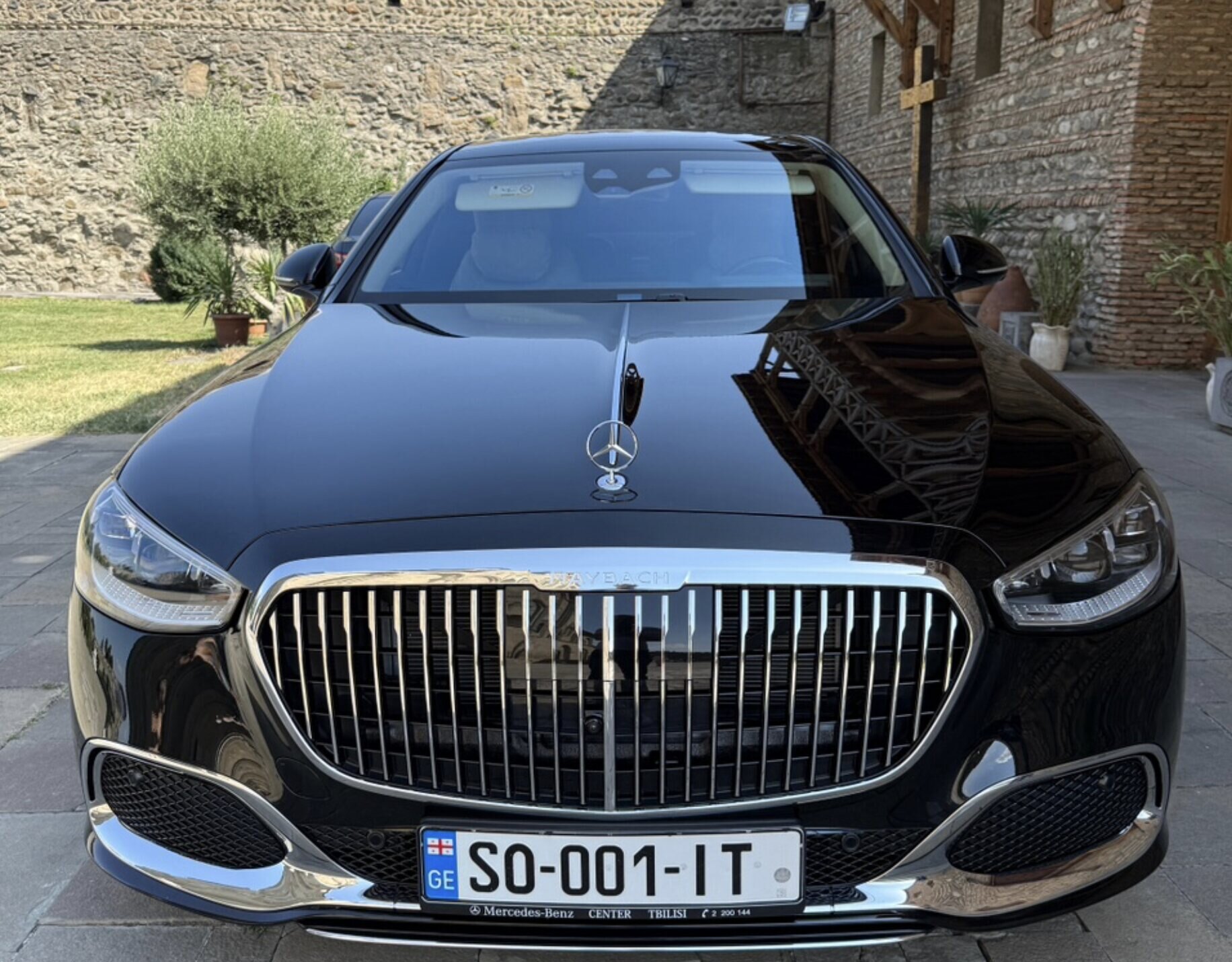 შავი Mercedes-Maybach წინიდან დანახული, ქვაფენილზე გაჩერებული ძველი ციხის კედელთან, დღის სინათლეზე.