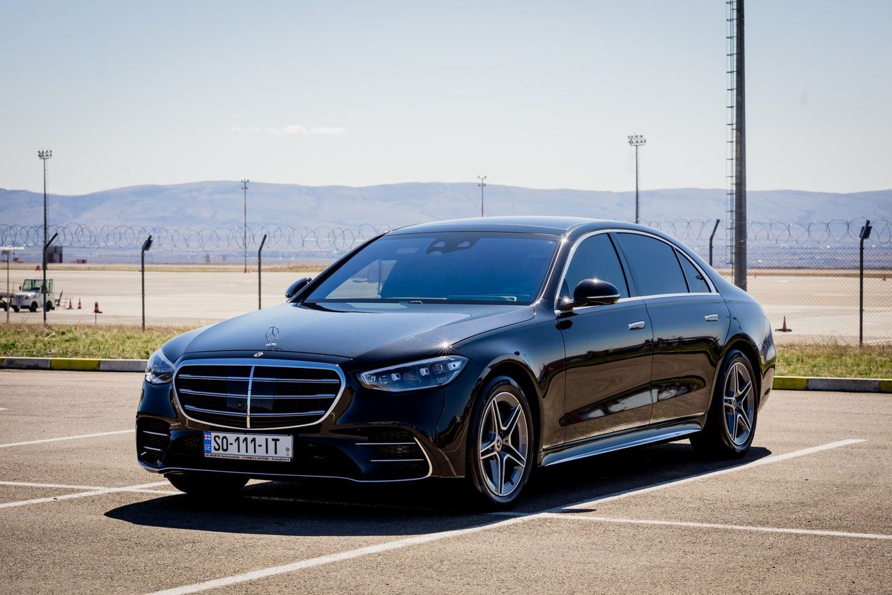 შავი Mercedes-Benz S-Class ქართული საფრენი ველის კიდეზე მოლოდინში, კავკასიონის წინამთები შორს ჩანს.