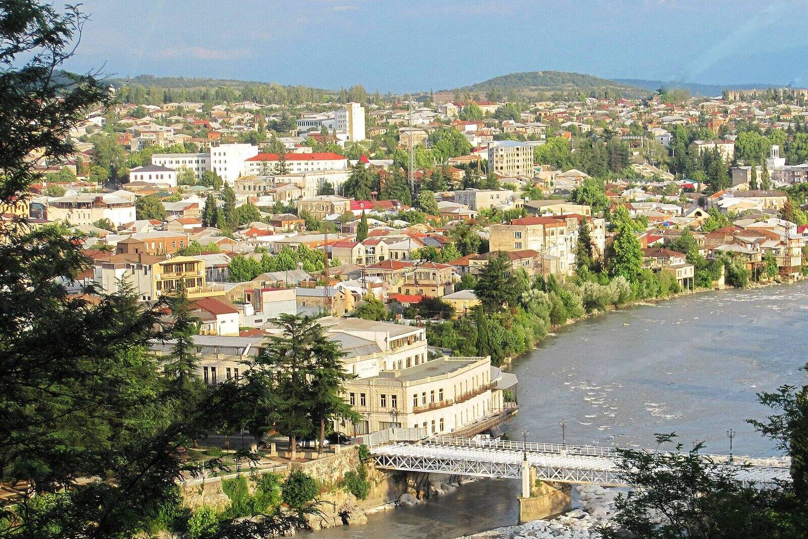 ქუთაისის ცენტრი ქალაქის ზემოთ მდებარე გორაკიდან: ქვემოთ რიონზე გადებული თეთრი ხიდი და სახურავები, რომლებიც მწვანე ბორცვებისკენ იშლება.