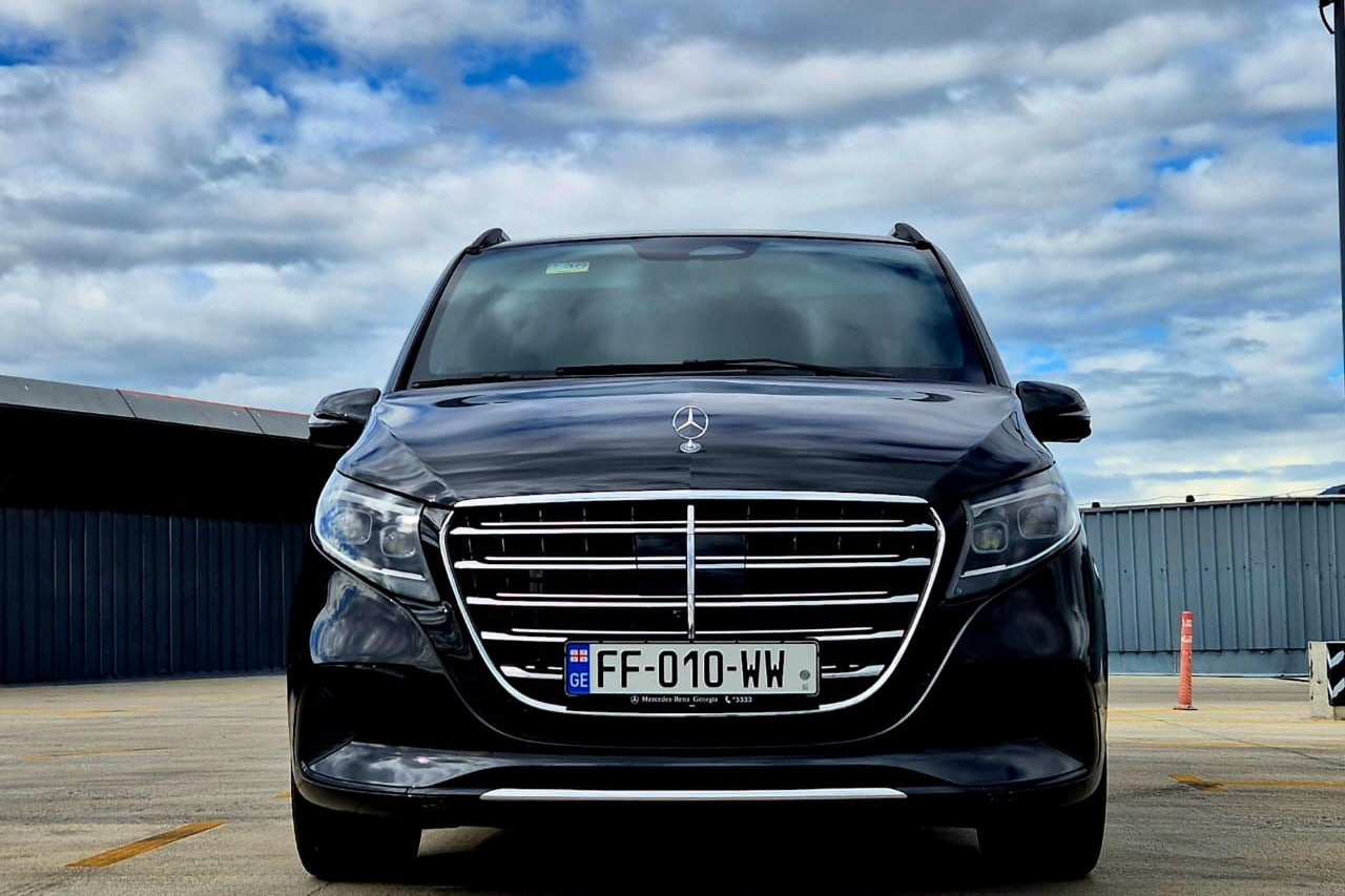 შავი Mercedes-Benz V-Class პირისპირ თბილისის საფრენ ველზე, ღრუბლები კაშკაშა ცის ფონზე.