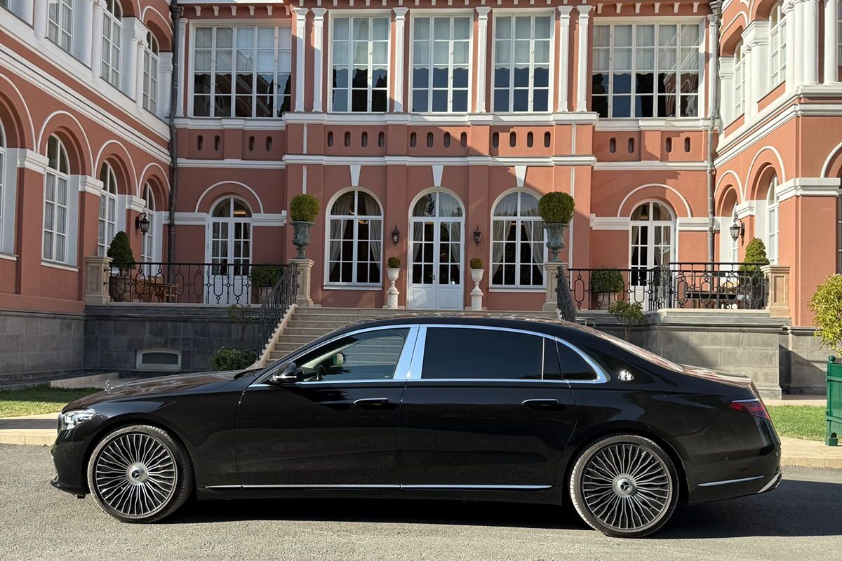 შავი Mercedes-Maybach S-Class გვერდულად დგას თაღოვანი ფანჯრებისა და ფართო ქვის კიბის მქონე წითელი აგურის სასახლის წინ.
